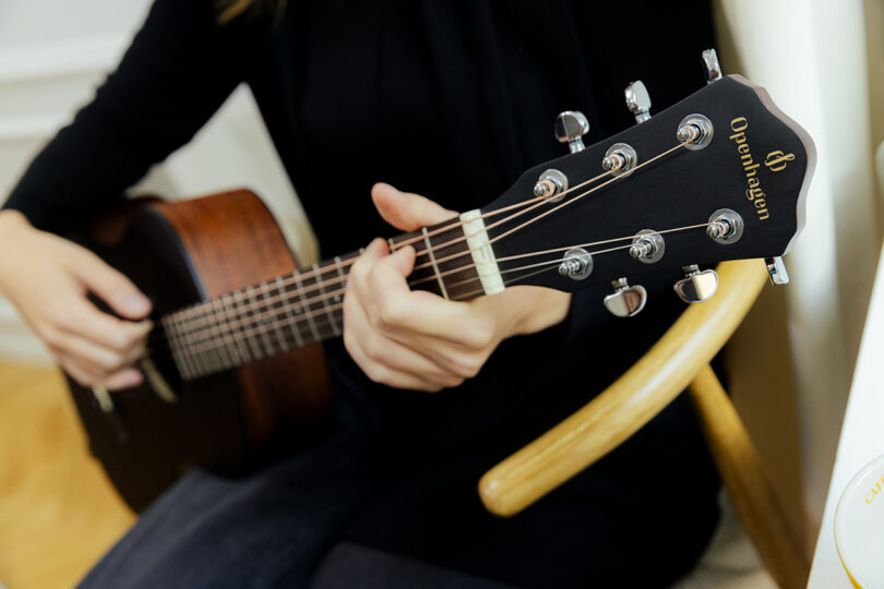 A person is seated playing an acoustic guitar, shown in close-up from the shoulders to the hands, focusing on the guitar's headstock and tuning pegs.