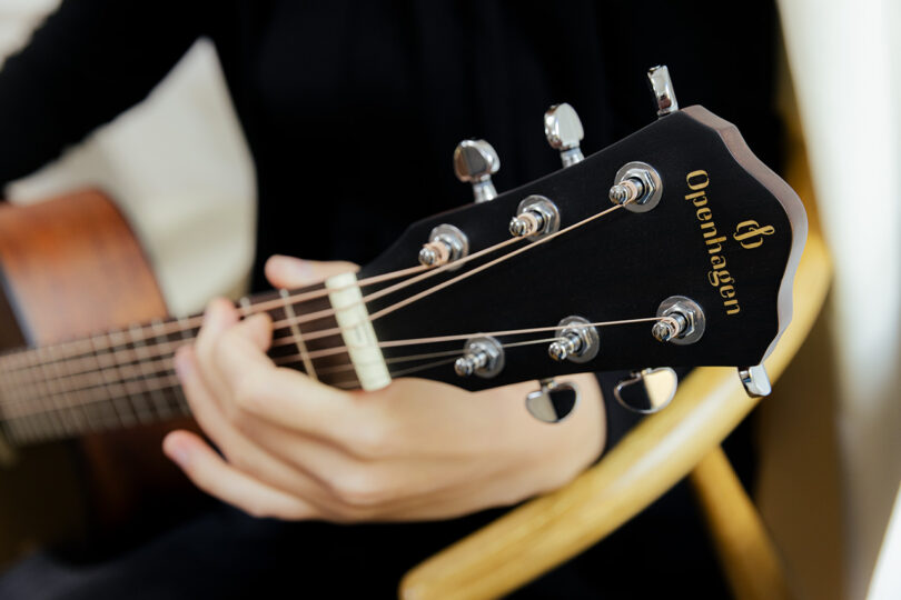 A person holding and playing an acoustic guitar with the brand name "Oppenhagen" visible on the headstock.
