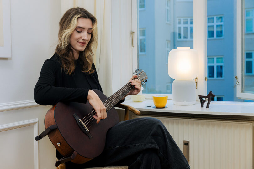 A woman sits indoors near a window, playing an acoustic guitar. A lamp, a yellow cup, and a small figurine are on the windowsill beside her.