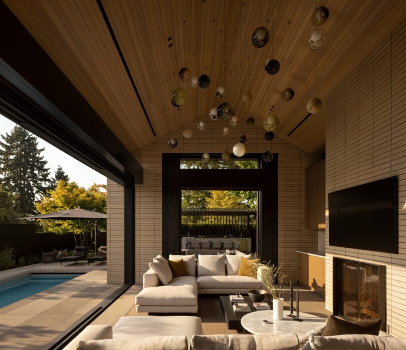 Modern indoor-outdoor living room with vaulted wood ceiling, neutral sofas, wall-mounted TV, fireplace, and view of a patio with pool and trees. Spherical light fixtures hang from the ceiling.