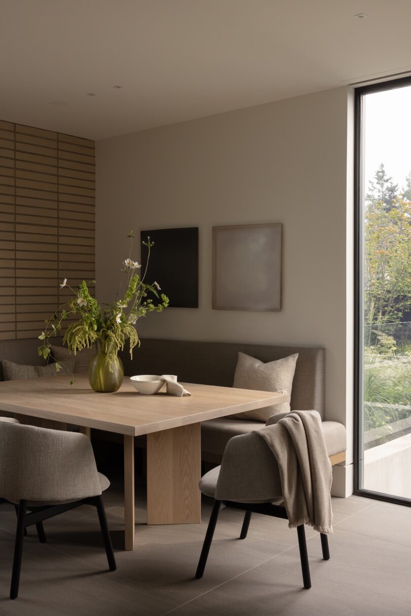 A modern dining area with a light wood table, beige chairs, a built-in bench, minimalist wall art, and a large window overlooking greenery. A vase with branches sits on the table.