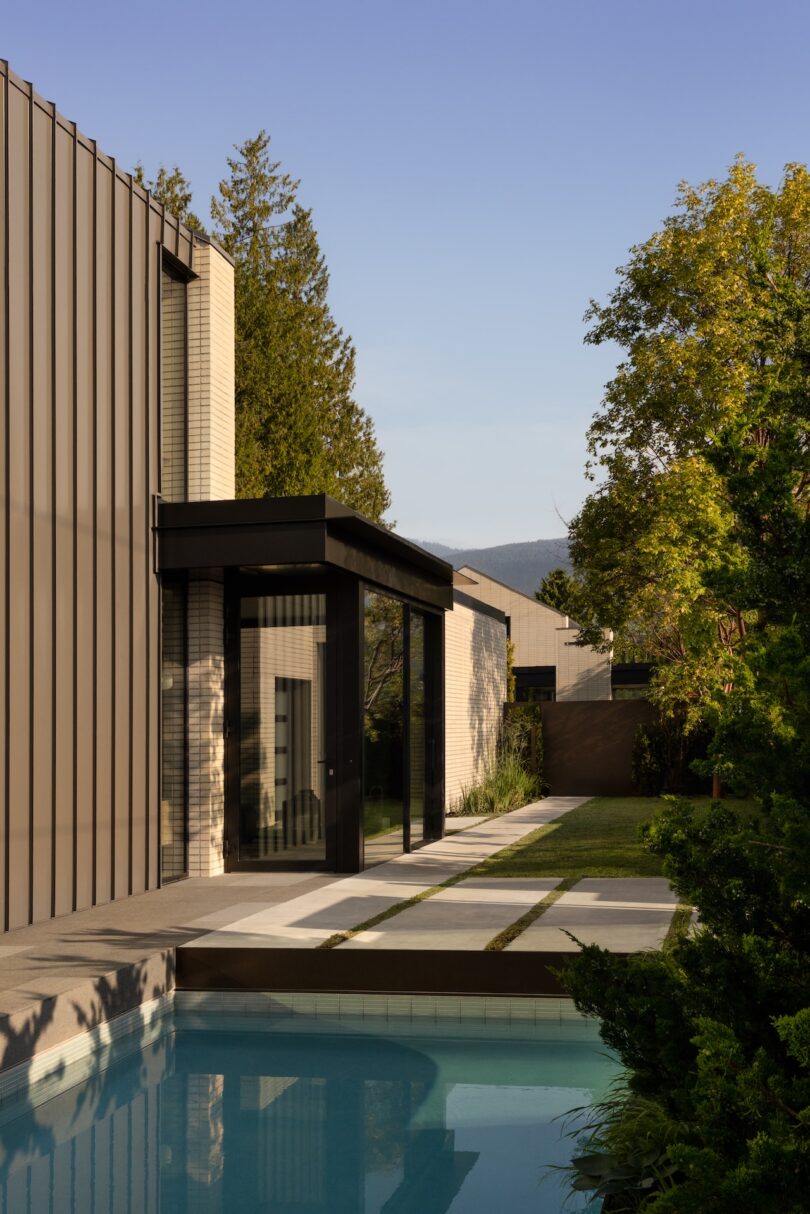 Modern house with vertical metal siding and large windows, adjacent to a rectangular swimming pool, surrounded by greenery and trees under a clear sky.