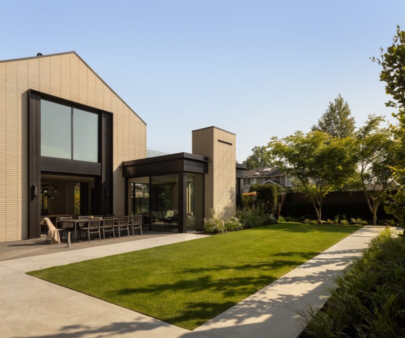 Modern house with large windows, outdoor dining area, and well-manicured lawn bordered by concrete paths and landscaped plants on a sunny day.