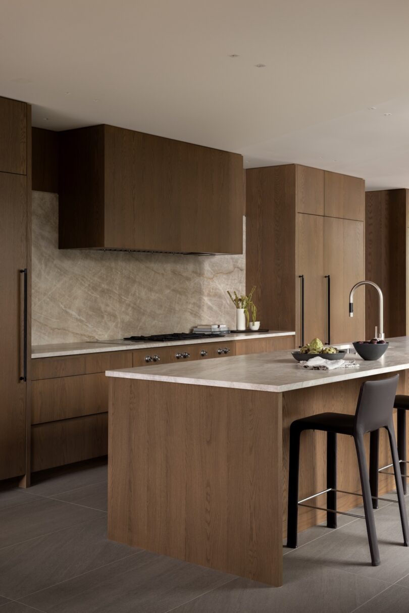 Modern kitchen with dark wood cabinets, a large island with gray countertop, built-in appliances, bar stool seating, and a marble backsplash.