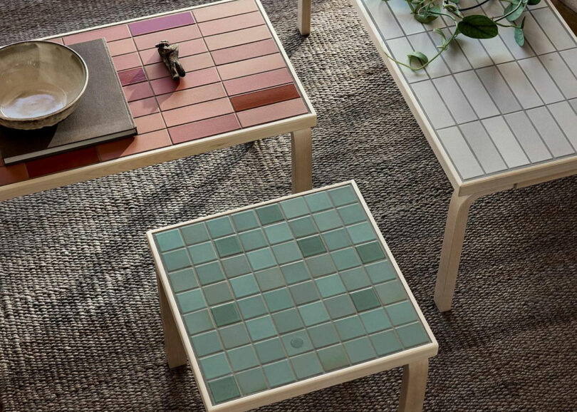 Three square Artek tables with tiled tops in red, cream, and green stand on a textured rug; one table has a plant and another holds a bowl and decorative object.