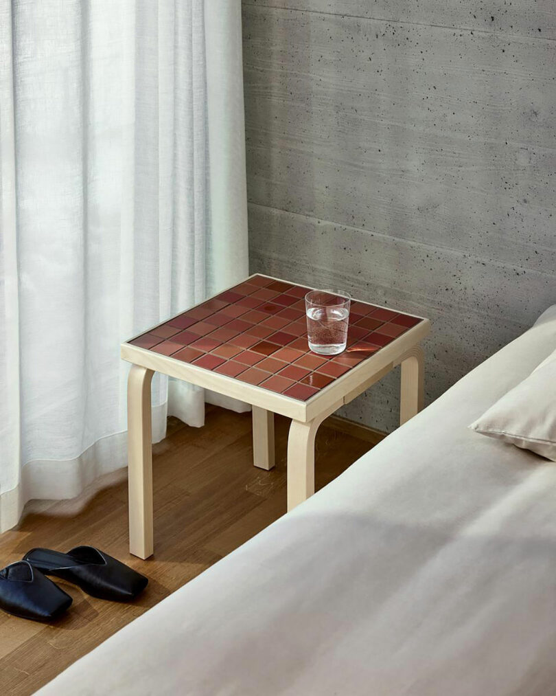 A small Artek bedside table with a red tiled top holds a glass of water; slippers are on the floor next to a bed in a minimalist room.