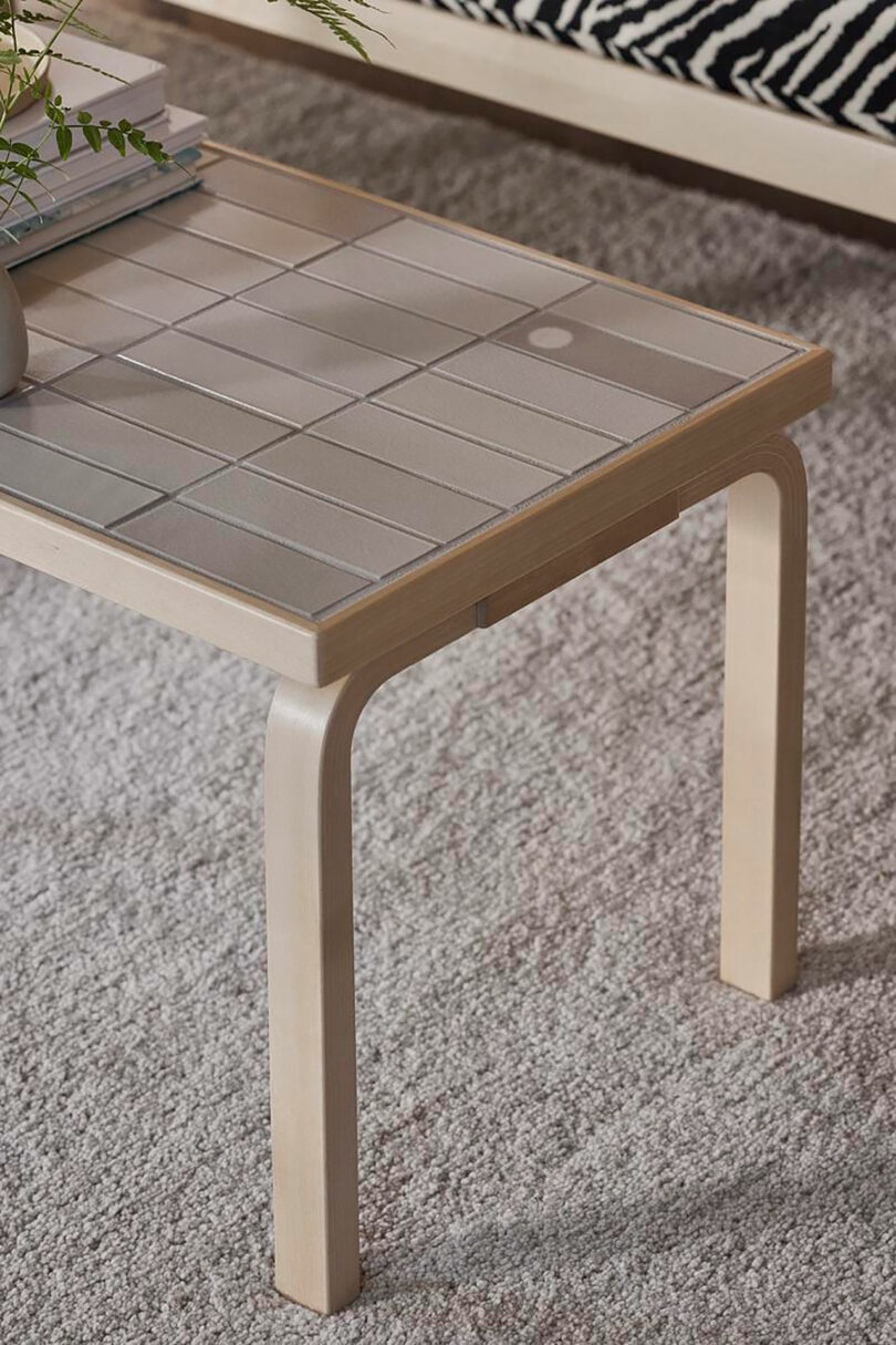 Rectangular Artek table with a tiled surface and light wood legs sits on a gray carpet, with books and a small plant on top.