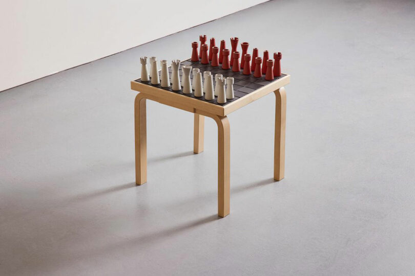 A small Artek wooden table with a built-in chessboard and neatly arranged white and red chess pieces stands on a plain gray floor.