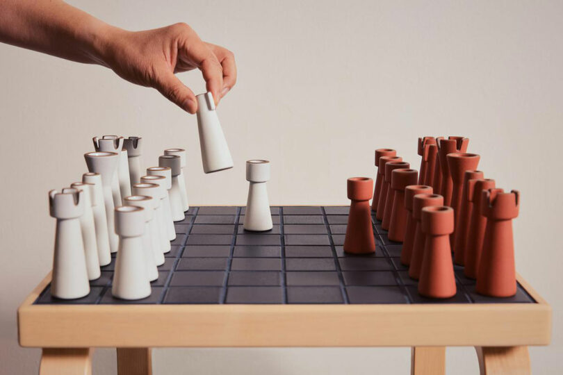 A hand moves a white chess piece on a minimalist Artek chess board adorned with elegant white and brown cone-shaped pieces.