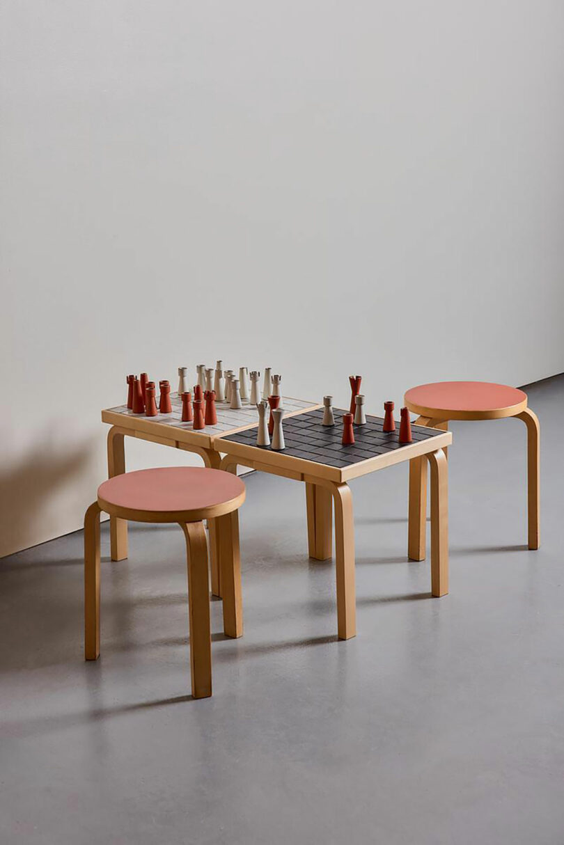 A minimalist chess set on a square wooden table, flanked by two round Artek wooden stools with reddish-orange seats, placed on a smooth grey floor against a plain wall.