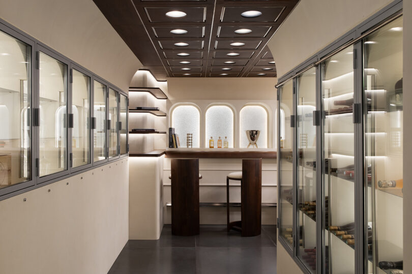 Modern wine tasting room with glass storage cabinets on both sides, a small bar area with two tall stools, shelves, and recessed lighting in the ceiling and walls.