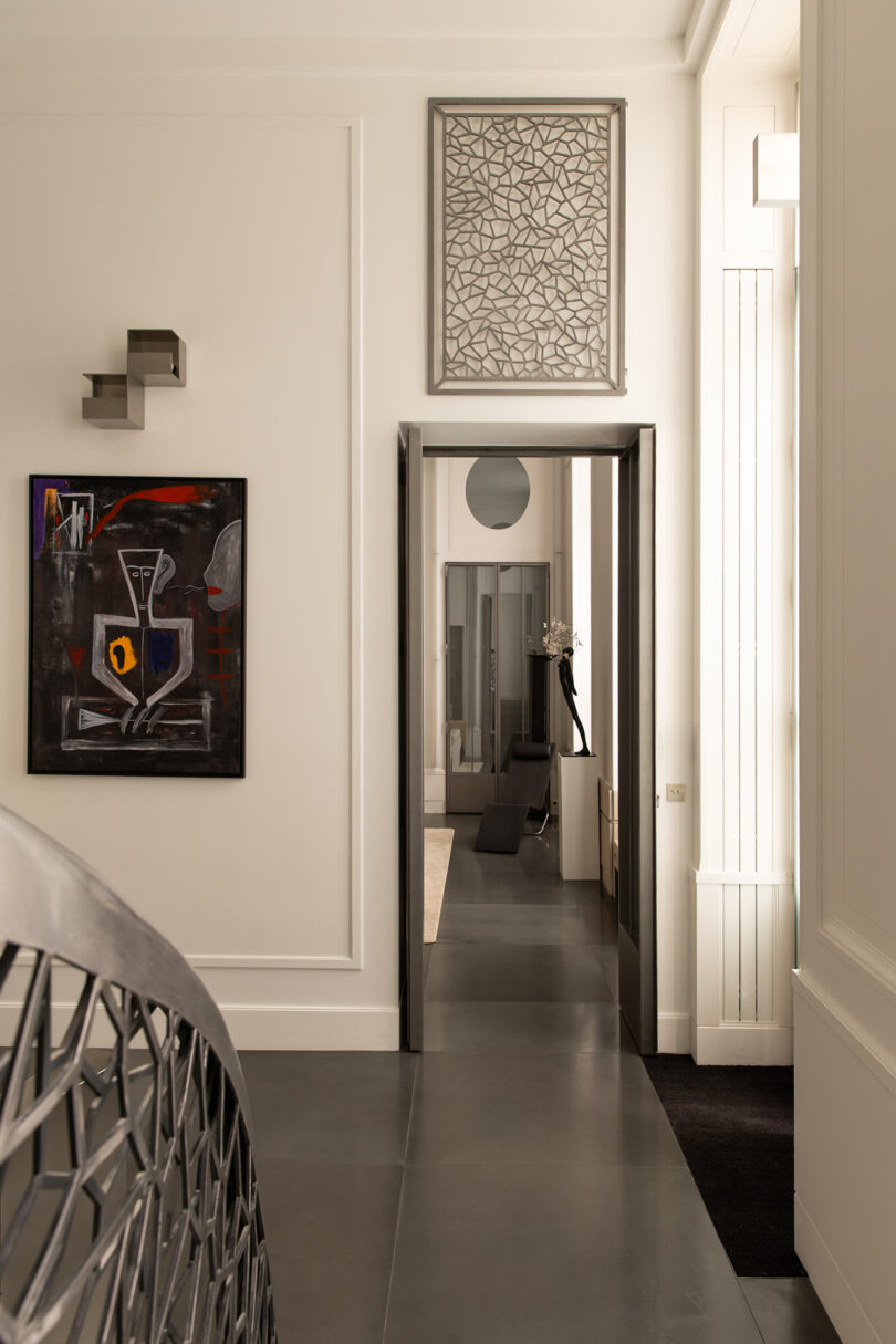 A modern hallway with geometric artwork, a black-and-red abstract painting, and sleek metal décor. The floor is dark, with minimal furnishings and neutral-toned walls.