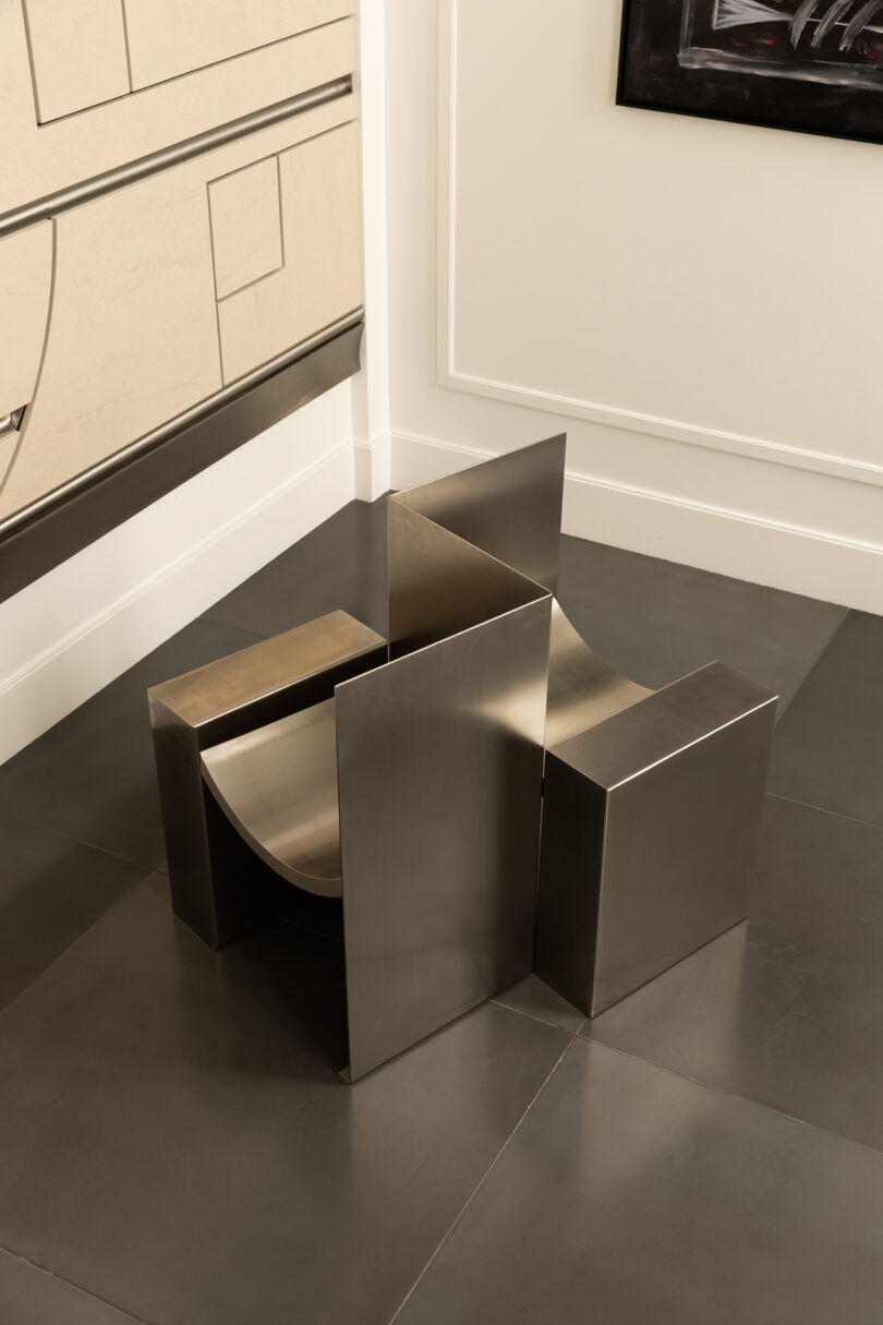 Modern metallic chair with geometric, angular design positioned on a tiled floor near a wall with minimalist art and trim.