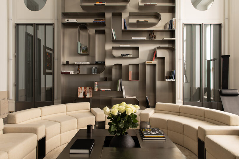 Modern living room with curved beige sofas, a black coffee table with books and flowers, and a large abstract wall bookshelf with various books and decorative items.