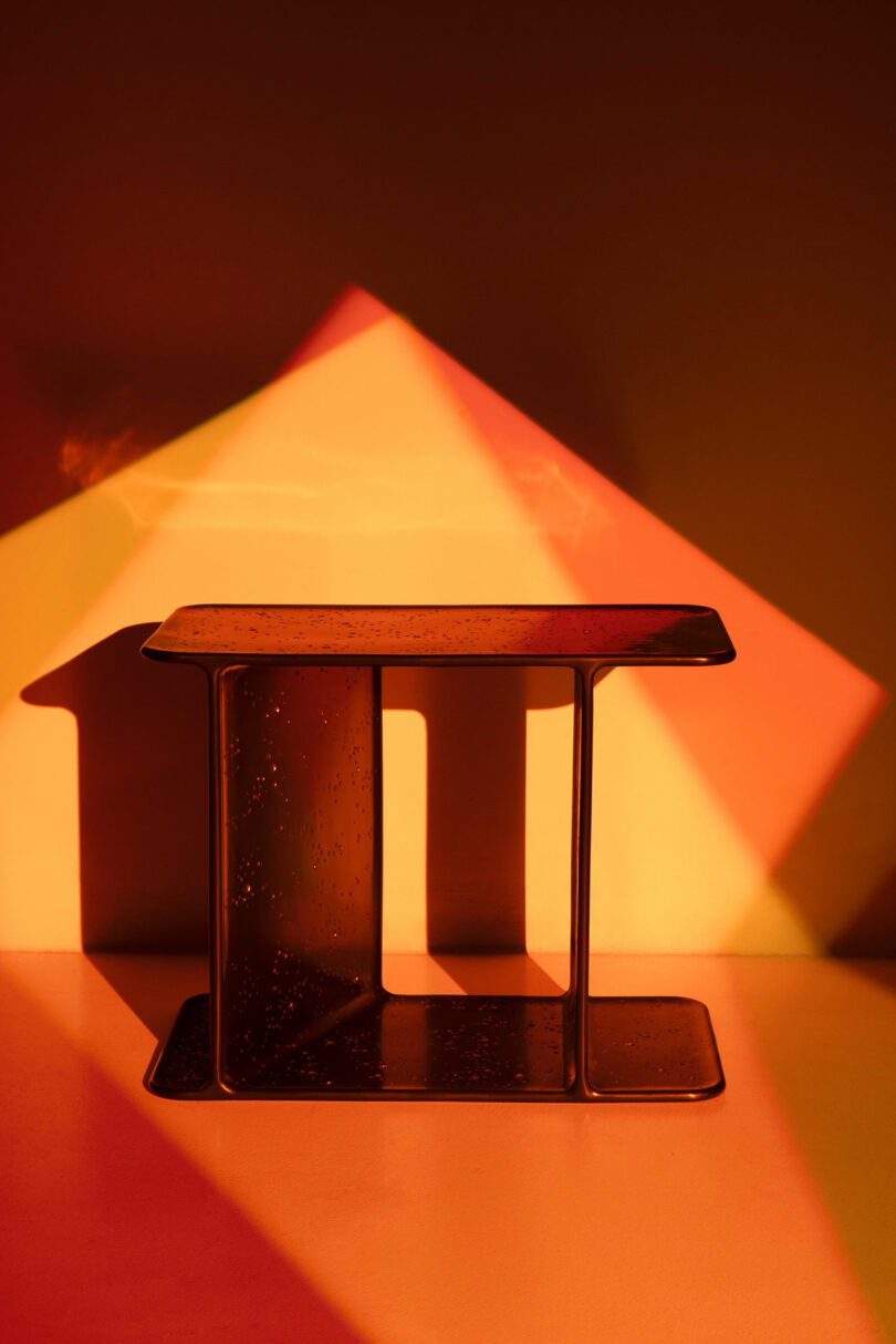A small, rectangular Holly Hunt metal table sits under warm, geometric light patterns casting sharp shadows on the wall and floor.