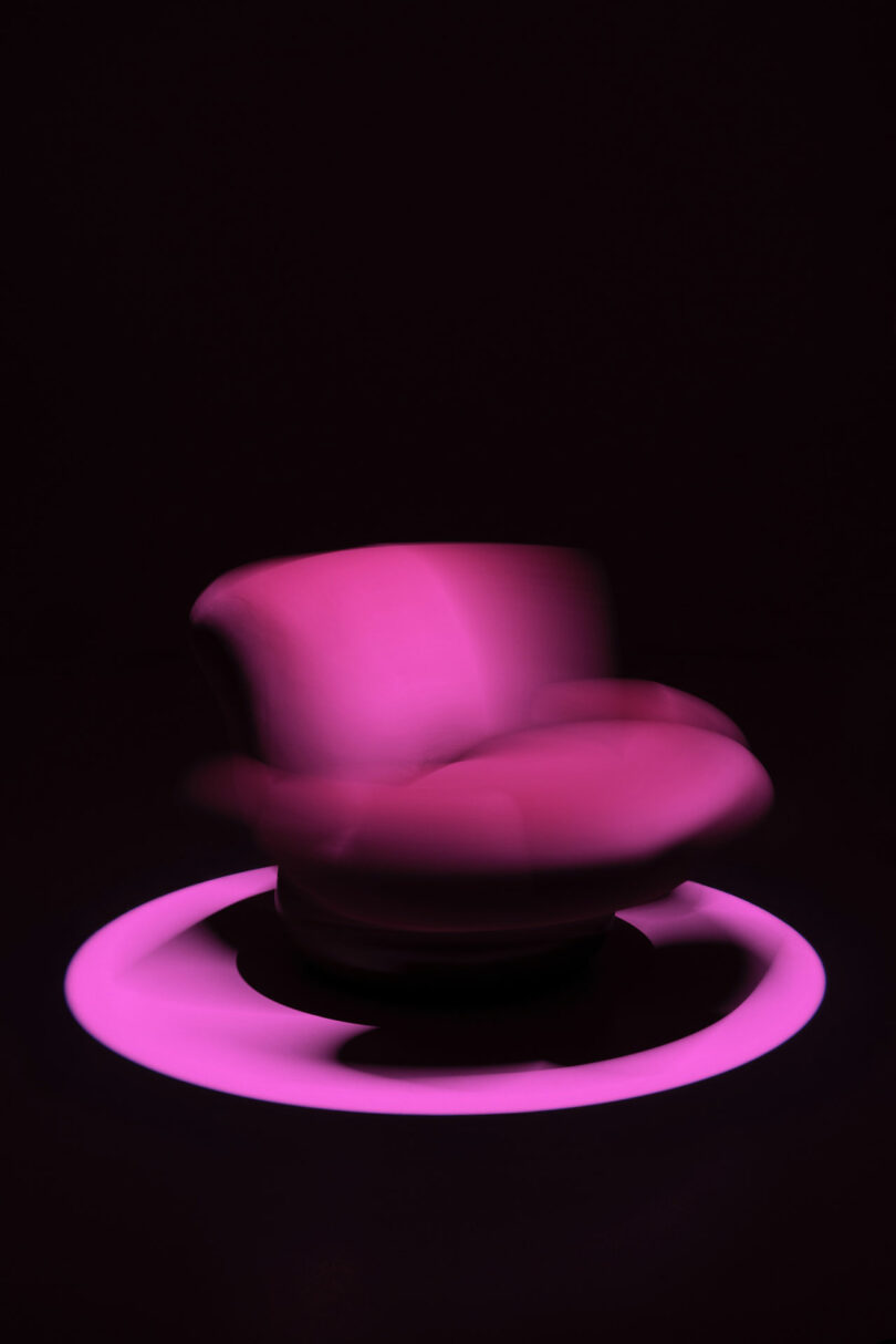 A Holly Hunt pink armchair sits in the center of a glowing pink ring against a dark background, with a slight blur suggesting motion.