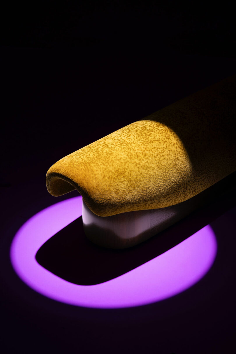 A close-up of a textured yellow object resembling a banana in a spotlight, accented by a purple ring of light on a dark surface—evoking the playful, modern aesthetic of Holly Hunt.