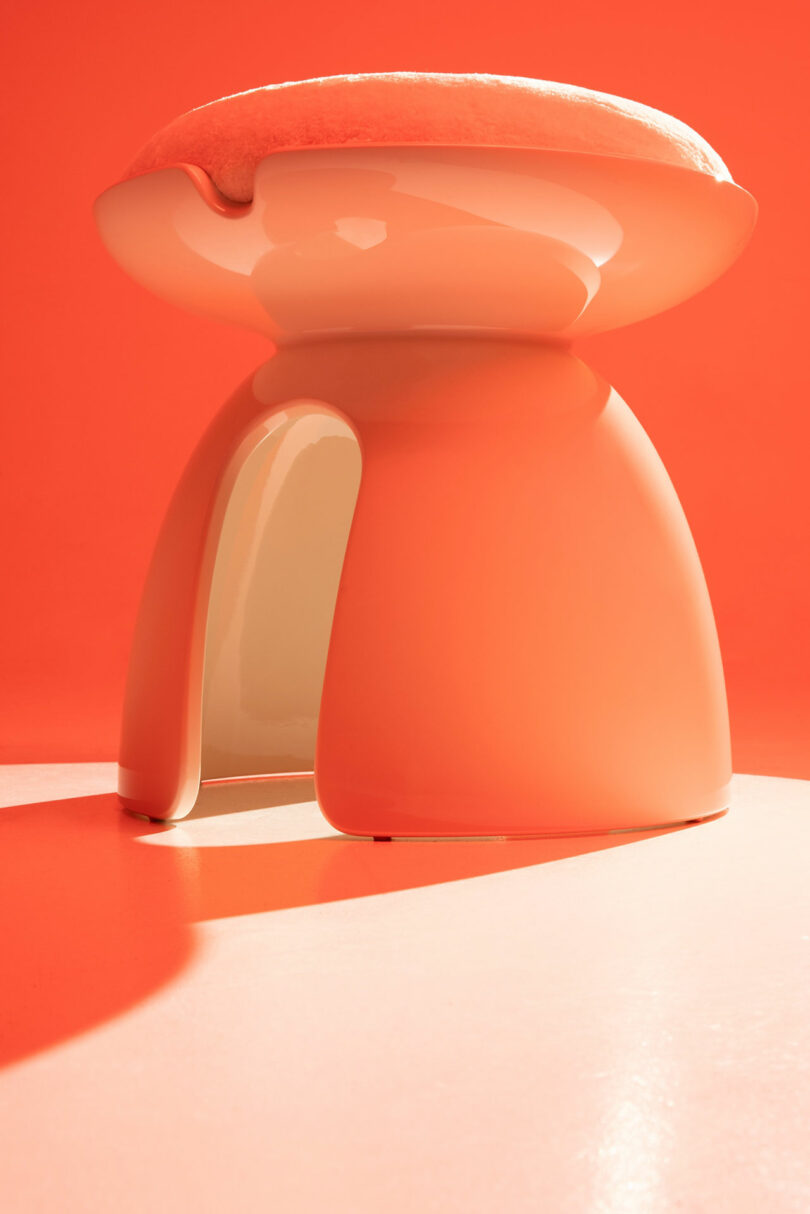 A glossy, modern orange stool by Holly Hunt, featuring a round cushioned top and an open side, sits on a matching orange surface against an orange background.