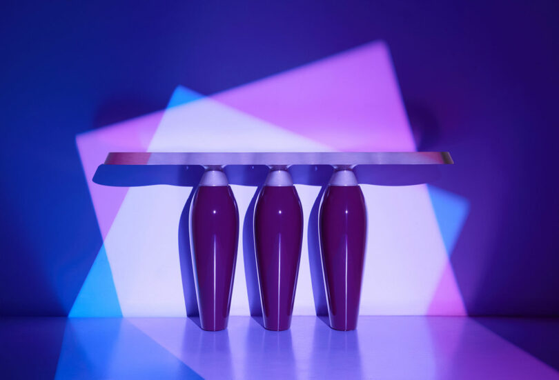 A modern Holly Hunt table with a rectangular top supported by three glossy purple cylindrical legs, illuminated by overlapping blue, purple, and pink lights.