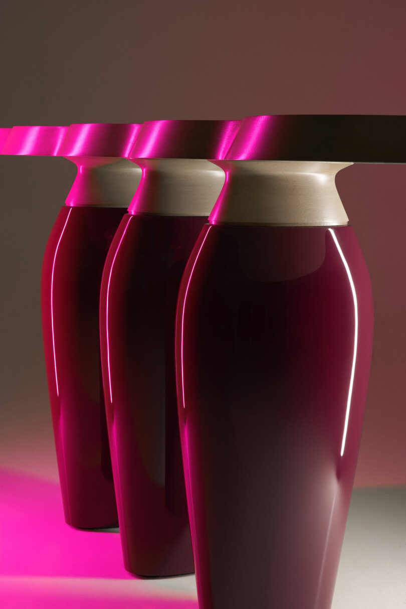 Three glossy, dark magenta vase-shaped table legs support a flat tabletop, reminiscent of Holly Hunt’s signature elegance, all illuminated by bright pink lighting from the left side.