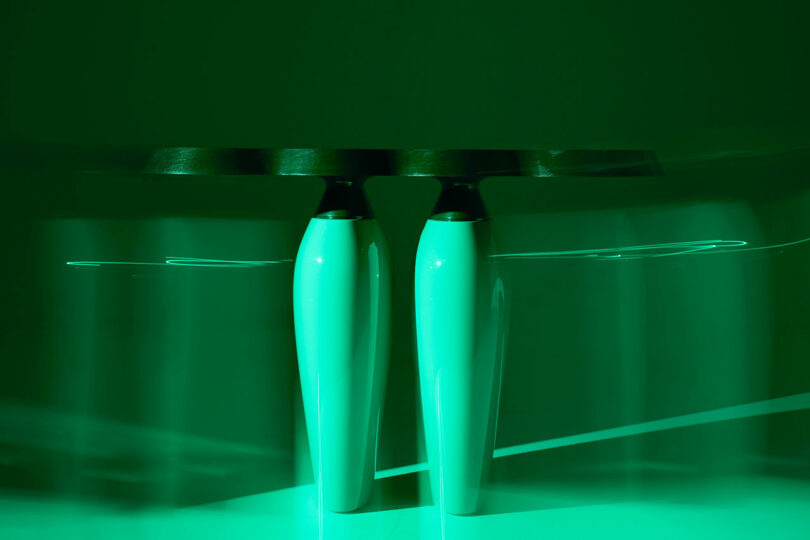 Two modern, glossy vases from Holly Hunt are placed side by side on a reflective surface, illuminated with strong green lighting and slight motion blur.