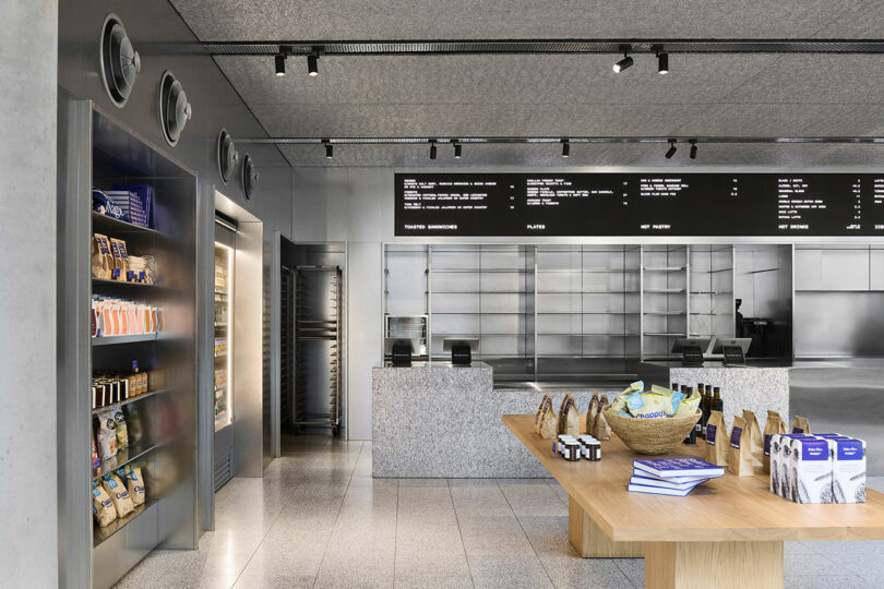 Modern bakery interior with metal shelves, a wooden display table with packaged goods, a refrigerated section, and digital menu boards above a granite counter.