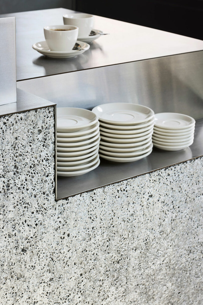Stacked white saucers and two white cups with saucers sit on a metallic countertop with a speckled textured front panel.