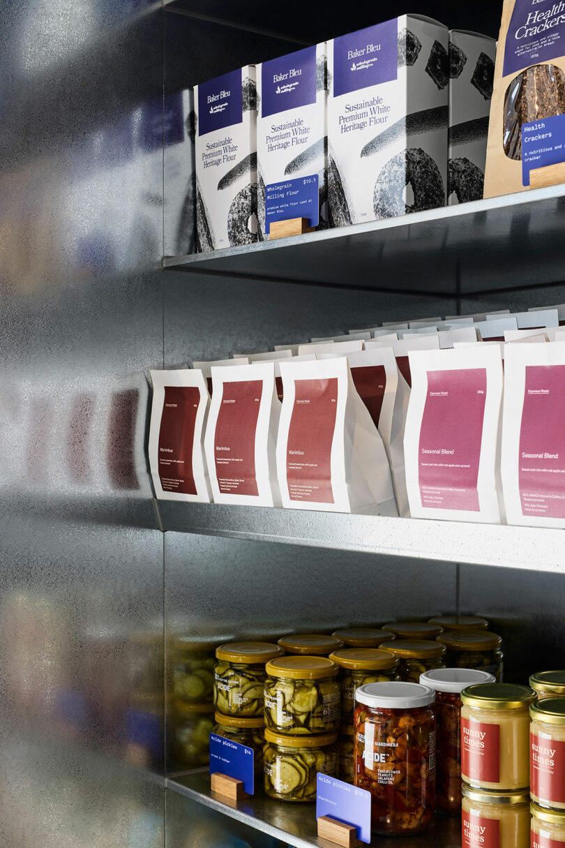 Shelves display packaged bread products, jars of pickles, and other preserved foods in a modern, metallic retail setting.