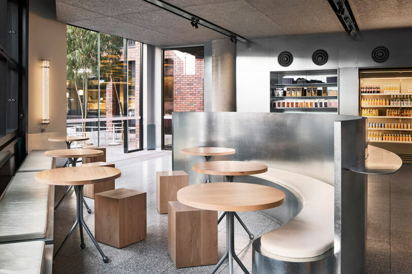 Modern café interior with round wooden tables, cube stools, a curved metal bench, large windows, and refrigerated shelves stocked with products in the background.