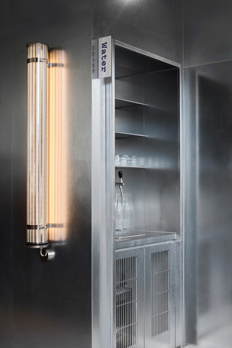 A metallic water station with a glass bottle and a faucet is set into a wall, next to a vertical, cylindrical light fixture. A sign labeled "Water" is visible above the station.