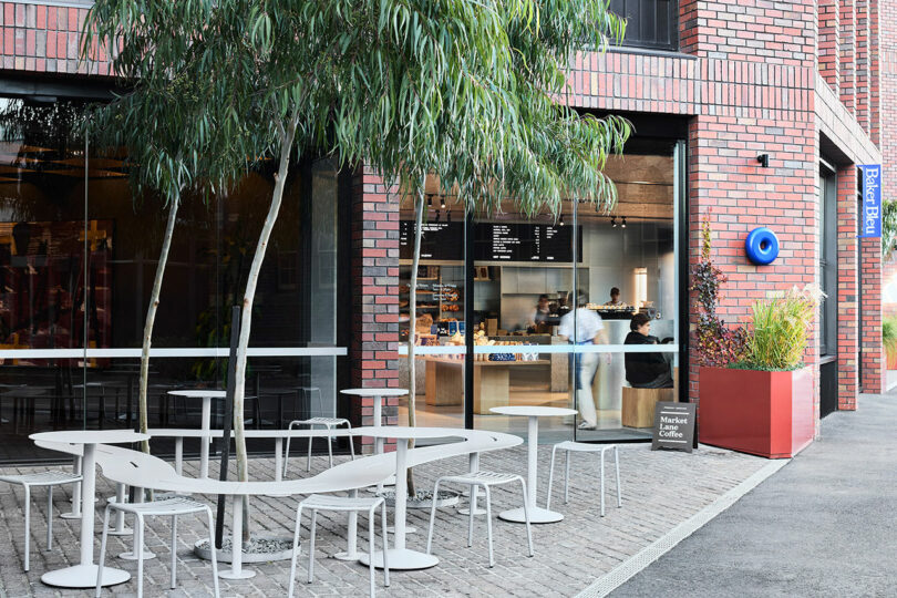 Outdoor seating area with white tables and chairs in front of a brick cafe; large windows reveal the interior counter and menu. A tree and large red planter are also visible.
