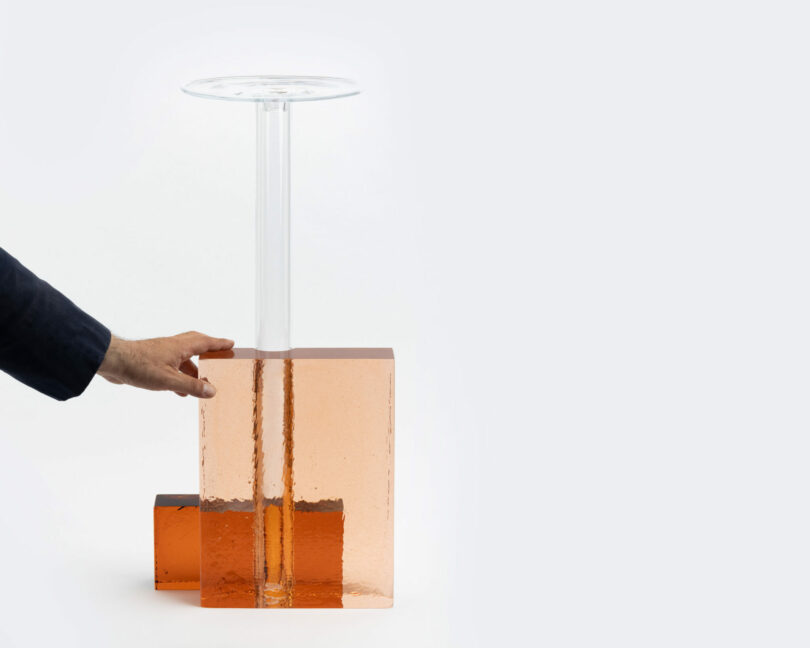 A hand touches a transparent orange block supporting a clear cylindrical column topped with a round, flat surface against a plain background.