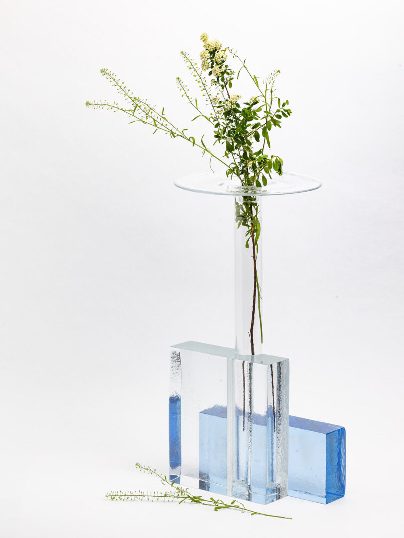 A clear glass vase with a single sprig of small white flowers stands on a round glass plate, next to two blue-tinted glass blocks; one sprig lies on the surface.