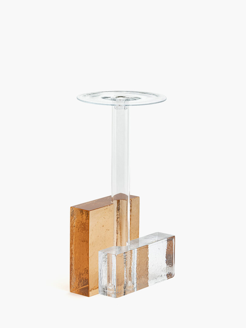 A modern glass sculpture with a clear vertical stem, a round top, and two rectangular bases—one amber and one clear.
