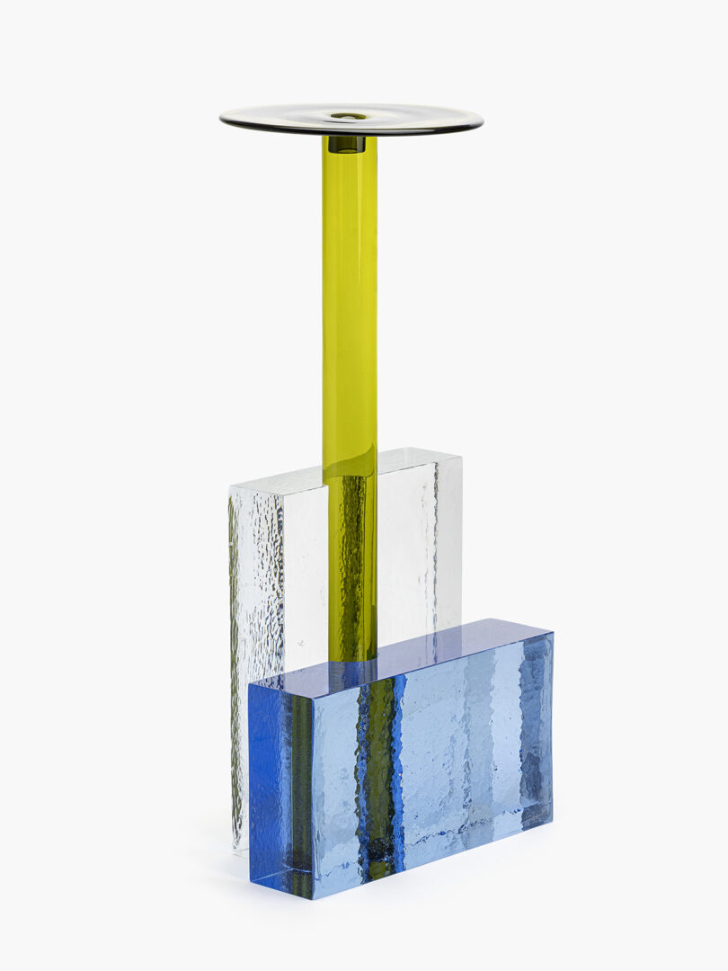 A modern form with a yellow cylindrical support, a round glass top, and rectangular blue and clear textured glass bases.