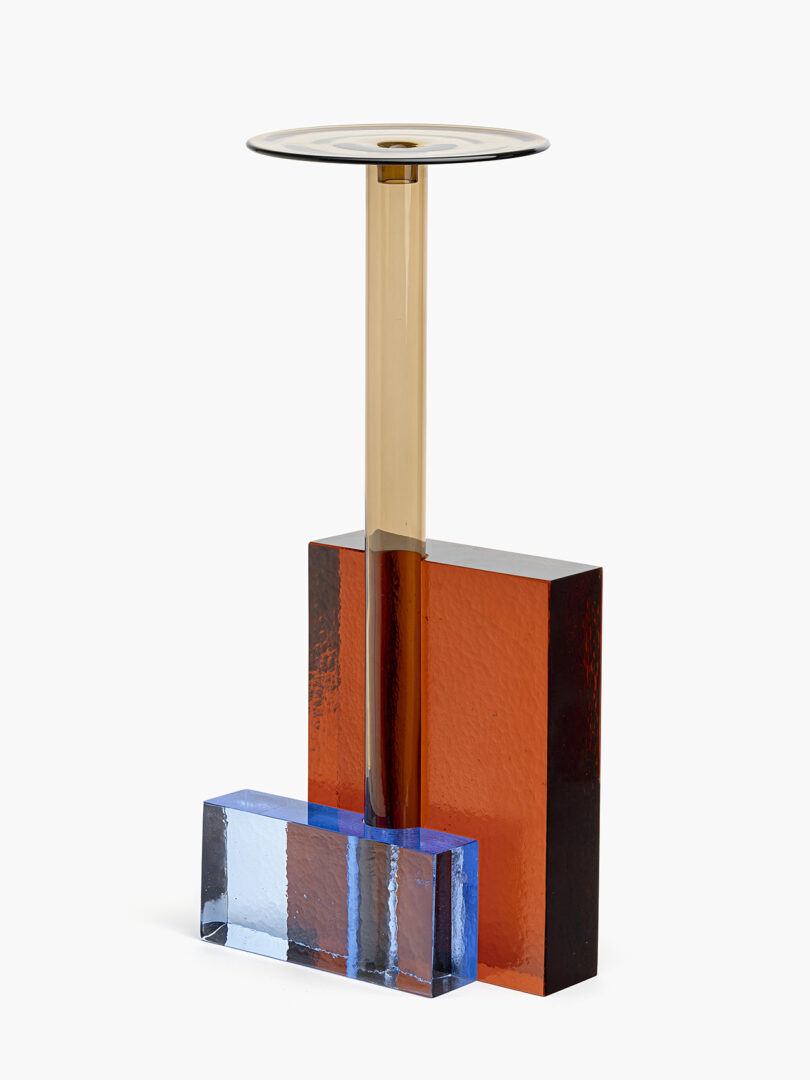 A modern sculpture with a gold cylindrical stand, red rectangular base, and blue rectangular accent, all made from glass or acrylic materials.
