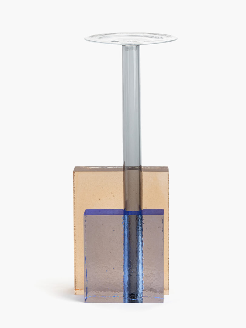 A modern form with a clear stem, a round transparent top, and a geometric base made of stacked rectangular glass blocks in amber and blue tones.