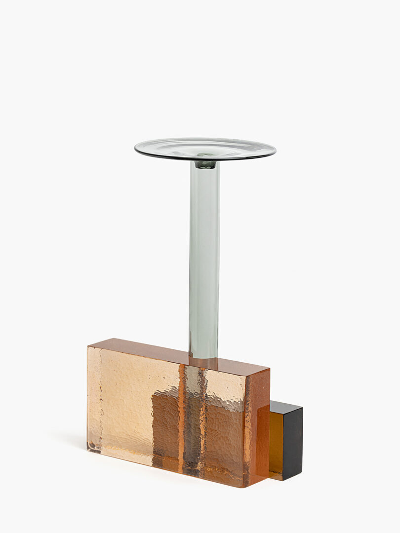 A modern form with a clear glass stem, a flat round top, and a rectangular amber-colored glass base with a small black cube at one end.
