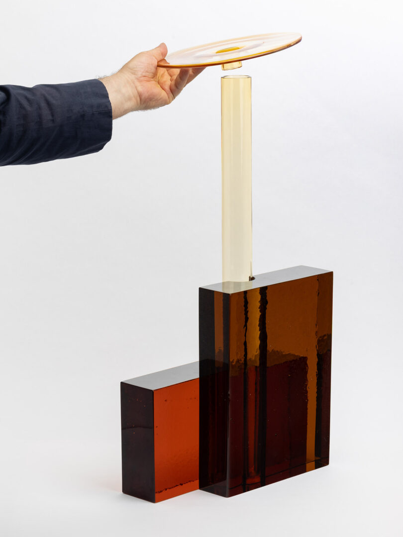 A hand places a translucent amber disc onto a vertical rod attached to two rectangular amber blocks of different heights, all made of glass or acrylic.