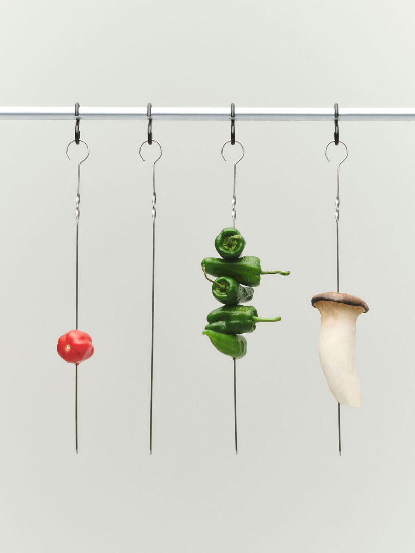 Four metal skewers with hooks hang from a rod; the skewers hold a red pepper, green peppers, and a king oyster mushroom, against a plain white background.