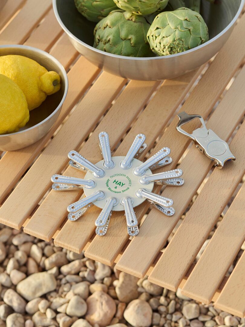 A metal table with bowls of artichokes and lemons, a bottle opener, and a circular clip holder, set on a wooden surface with rocks below.