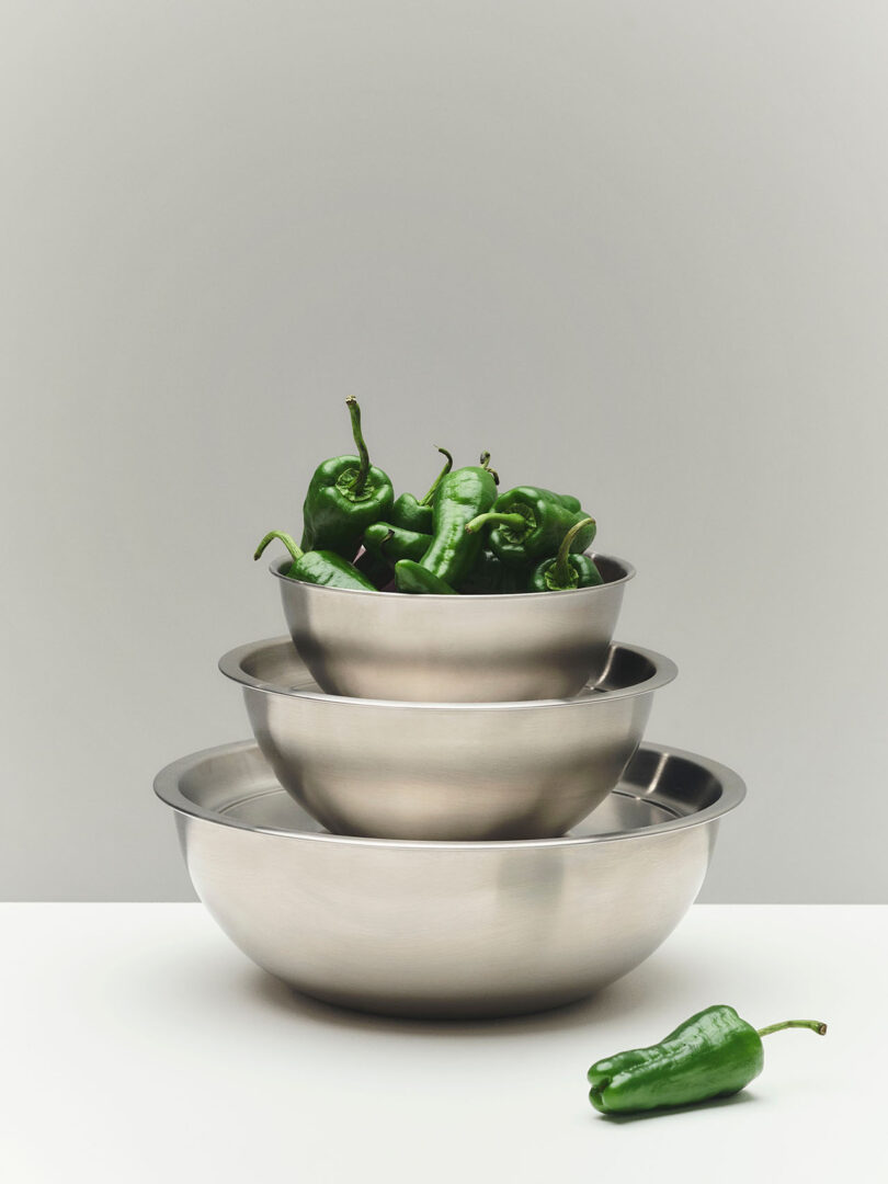 Three stainless steel mixing bowls stacked on a white surface, with the top bowl filled with green peppers and one pepper lying beside the bowls.