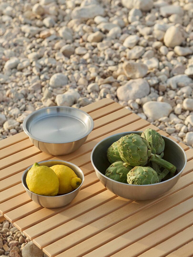 Three metal bowls sit on a wooden slatted surface, containing artichokes, lemons, and a covered bowl, with rocks visible in the background.