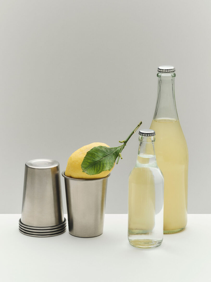 Two metal cups, one holding a lemon with a leaf, are beside a stack of cups. In front are two glass bottles, one filled with clear liquid and one with pale yellow liquid.
