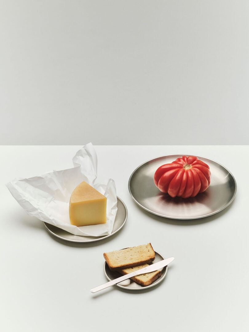 A plate with a large heirloom tomato, a plate with a wedge of cheese on parchment, and a small plate with two slices of bread and a butter knife on a white surface.