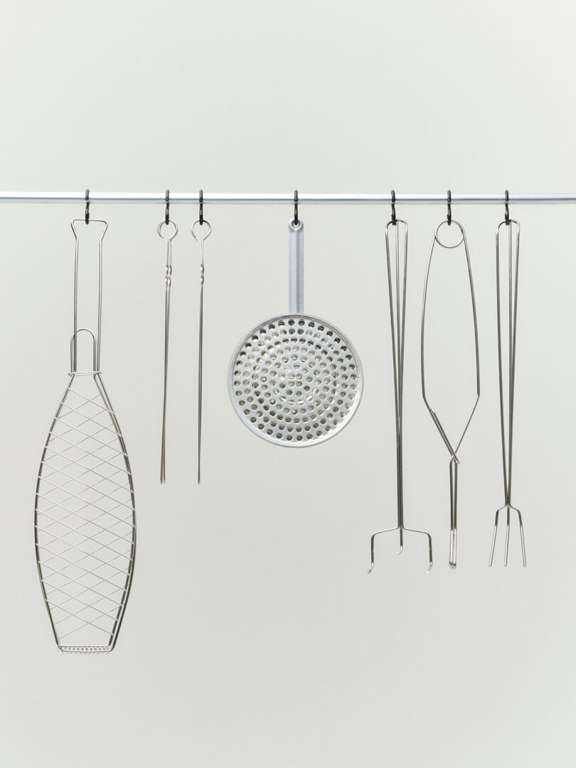 Six metal barbecue tools, including skewers, skewered forks, a grill basket, and a round grill grate, hang from hooks on a metal bar against a plain light background.