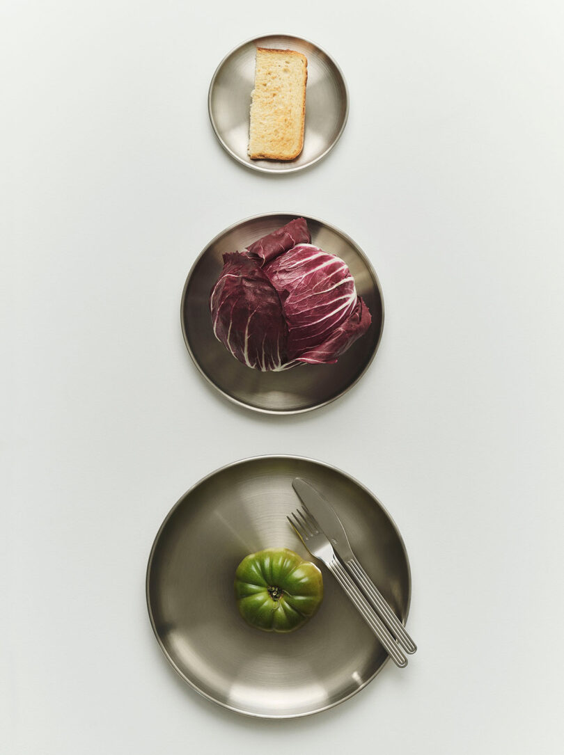 Three metal plates are arranged vertically: the top plate has a slice of bread, the middle has a head of radicchio, and the bottom has a green tomato with a fork and knife.