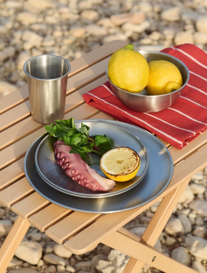 A metal plate with a cooked octopus tentacle, grilled lemon half, and parsley on a wooden table, next to a metal cup, a bowl of lemons, and a red striped cloth.