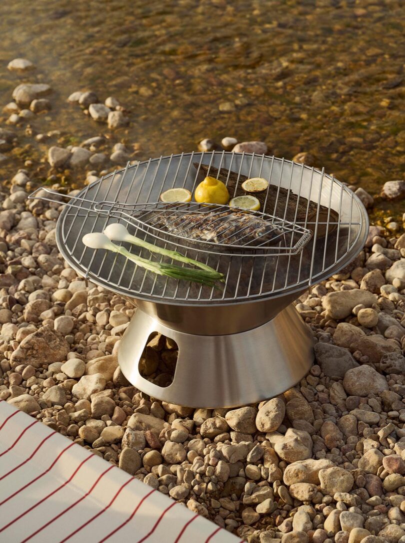A portable metal grill with food including scallions, lemon halves, and what appears to be fish, set on a rocky riverbank near the water.