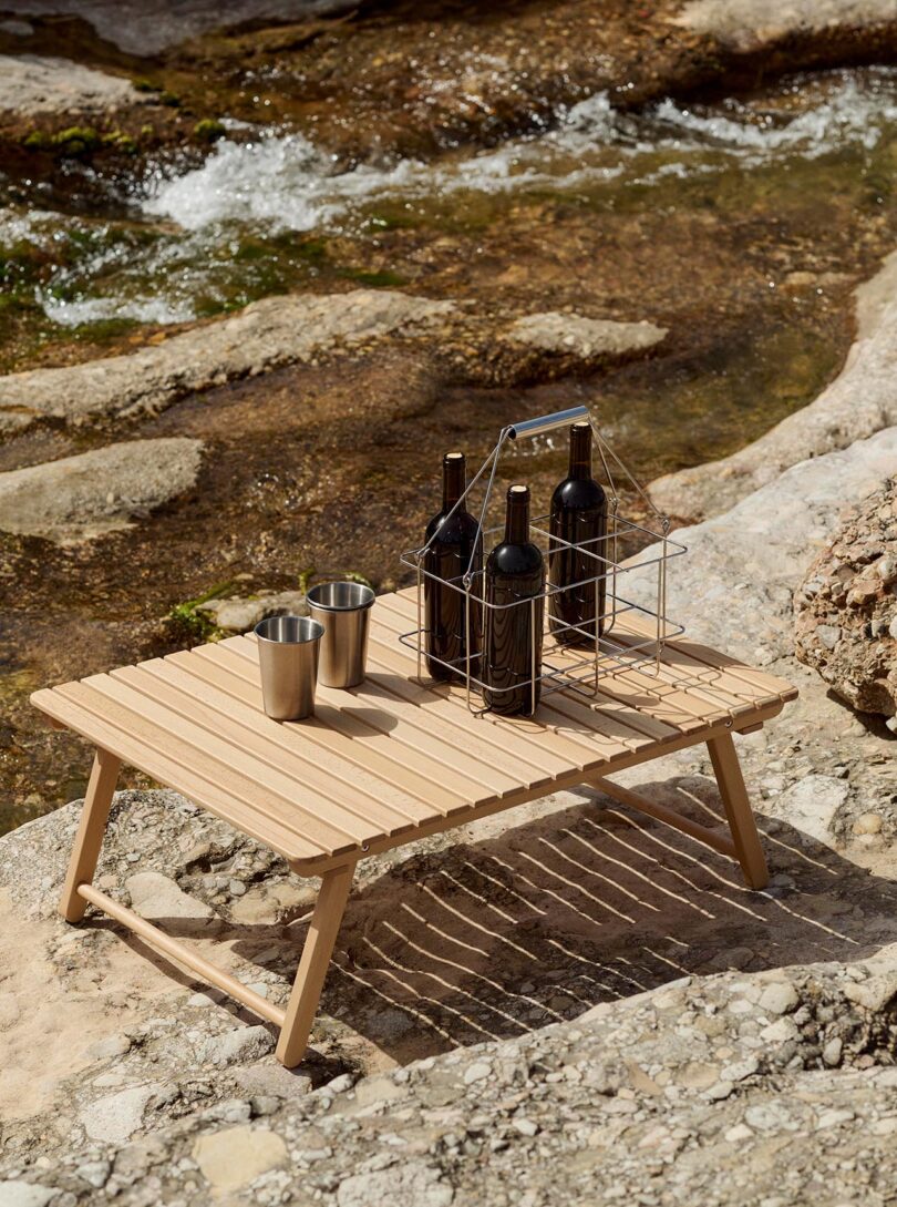 A wooden picnic table with metal cups and a metal carrier holding four bottles of wine, set on rocky terrain beside a flowing stream.